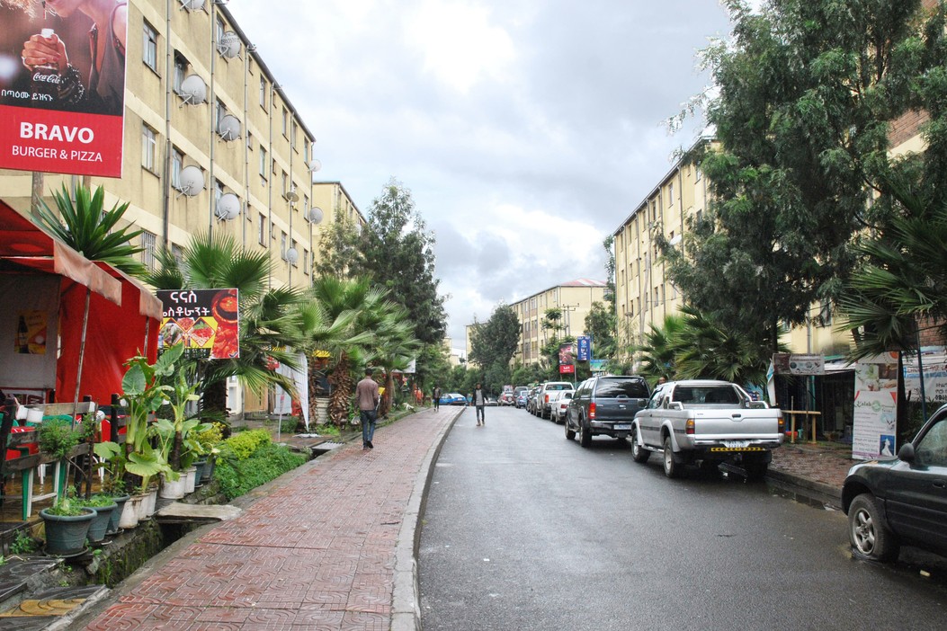 A tree-lined street in Addis Ababa on an overcast day. Cars line the street on the right. On the left is a restaurant patio.